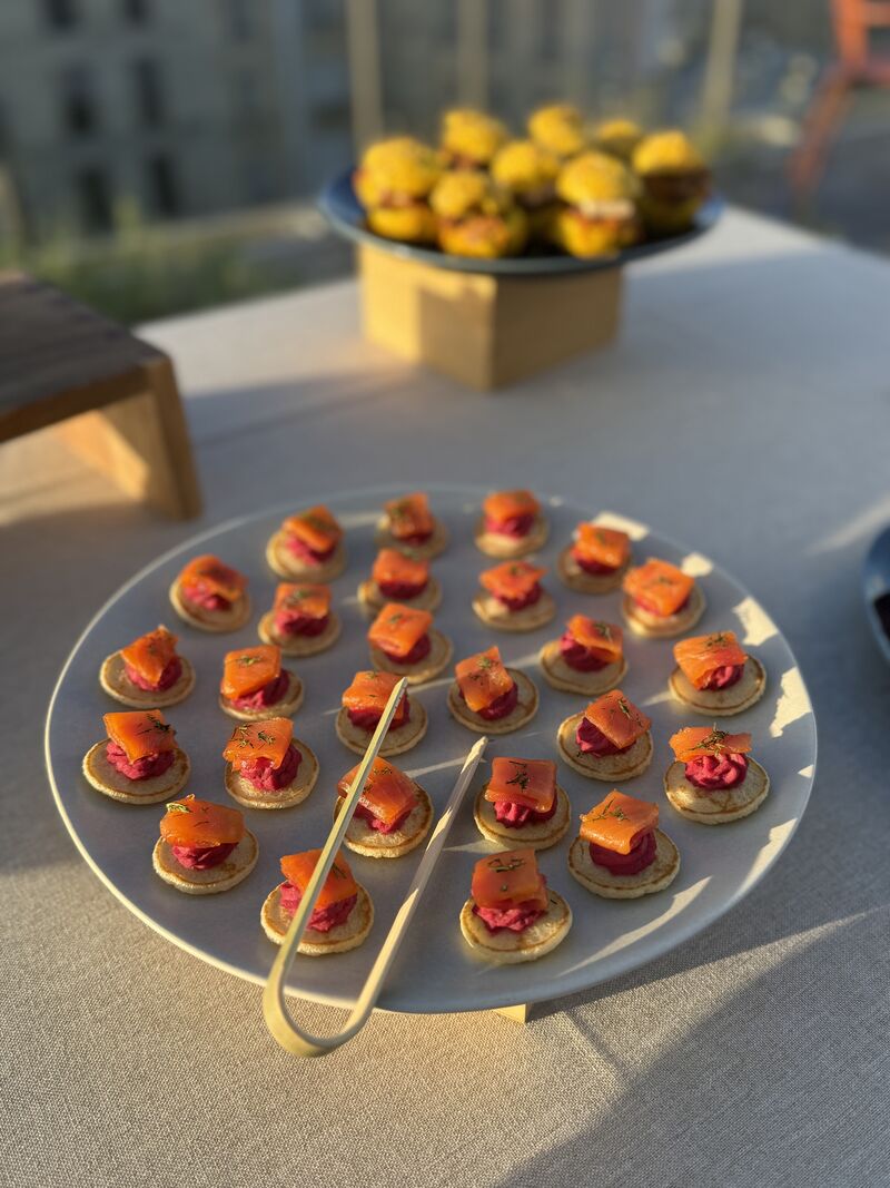 Une cheffe souriante en tenue blanche dresse une table élégante en extérieur avec des amuse-bouches, des plats colorés et des compositions florales, illustrant une expérience traiteur printanière raffinée et conviviale.