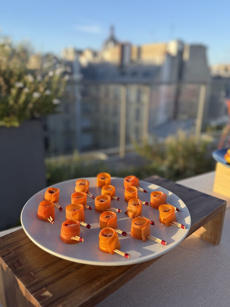 Une main verse une boisson rose rafraîchissante dans un verre sur une table élégante en extérieur, entourée de fruits, de fleurs et de plats colorés, illustrant une ambiance conviviale et estivale.