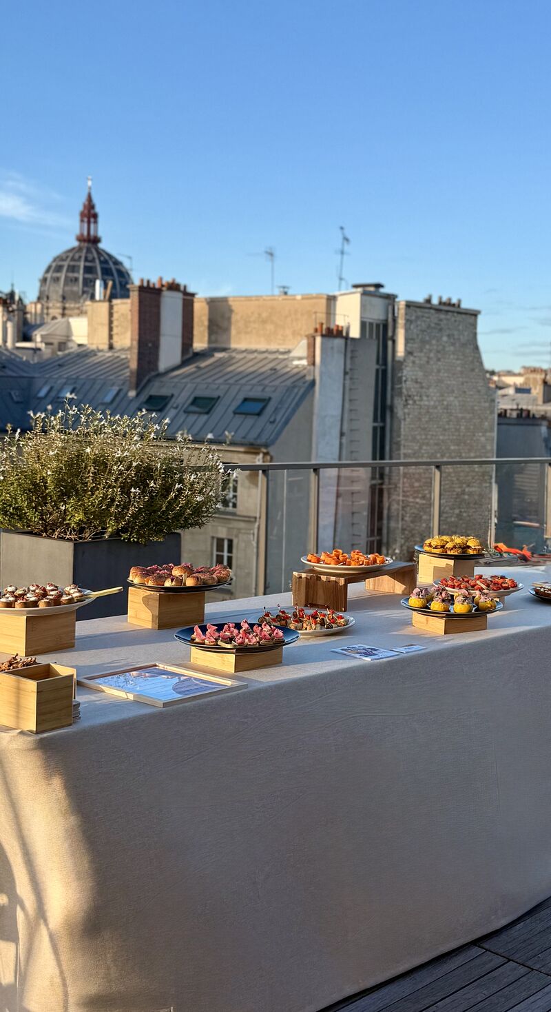 Une table extérieure élégante garnie de fruits frais, légumes de saison, pastèque, agrumes et bouchées apéritives, accompagnée de compositions florales colorées, illustrant une expérience traiteur printanière fraîche et conviviale.