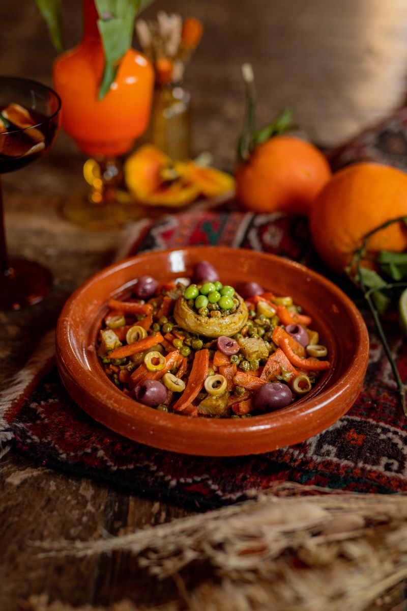 Une cheffe souriante en tenue blanche dresse une table élégante en extérieur avec des amuse-bouches, des plats colorés et des compositions florales, illustrant une expérience traiteur printanière raffinée et conviviale.