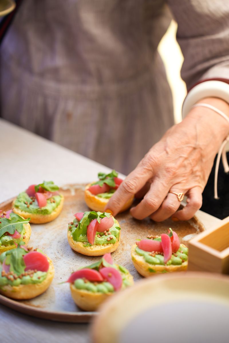 Une cheffe souriante en tenue blanche dresse une table élégante en extérieur avec des amuse-bouches, des plats colorés et des compositions florales, illustrant une expérience traiteur printanière raffinée et conviviale.