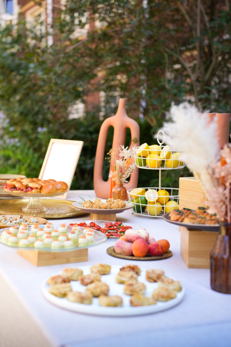 Une table extérieure élégante garnie de fruits frais, légumes de saison, pastèque, agrumes et bouchées apéritives, accompagnée de compositions florales colorées, illustrant une expérience traiteur printanière fraîche et conviviale.