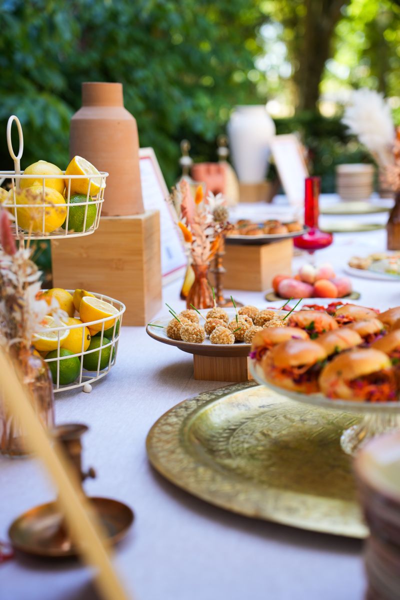 Une main verse une boisson rose rafraîchissante dans un verre sur une table élégante en extérieur, entourée de fruits, de fleurs et de plats colorés, illustrant une ambiance conviviale et estivale.