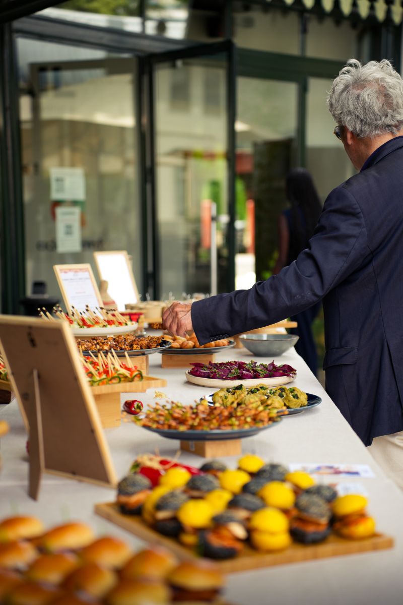 Une table extérieure élégante garnie de fruits frais, légumes de saison, pastèque, agrumes et bouchées apéritives, accompagnée de compositions florales colorées, illustrant une expérience traiteur printanière fraîche et conviviale.
