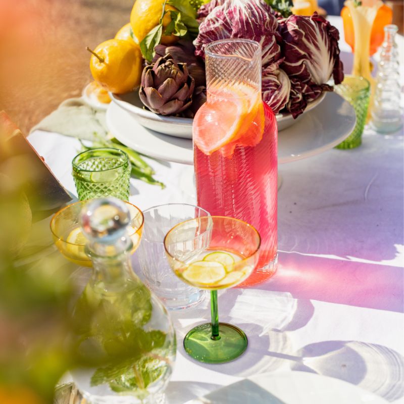 Une table extérieure élégante garnie de fruits frais, légumes de saison, pastèque, agrumes et bouchées apéritives, accompagnée de compositions florales colorées, illustrant une expérience traiteur printanière fraîche et conviviale.