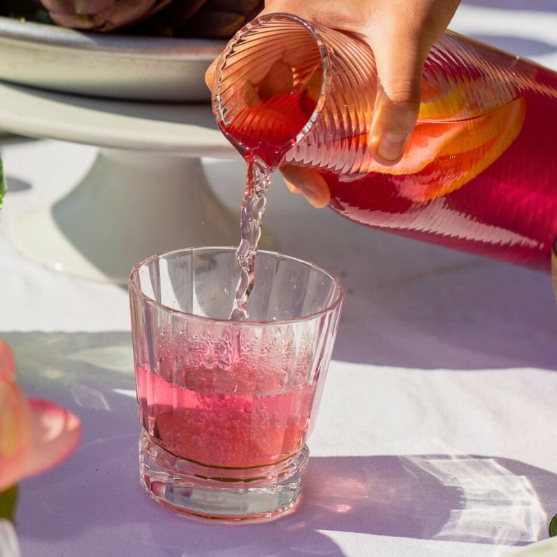 Une main verse une boisson rose rafraîchissante dans un verre sur une table élégante en extérieur, entourée de fruits, de fleurs et de plats colorés, illustrant une ambiance conviviale et estivale.