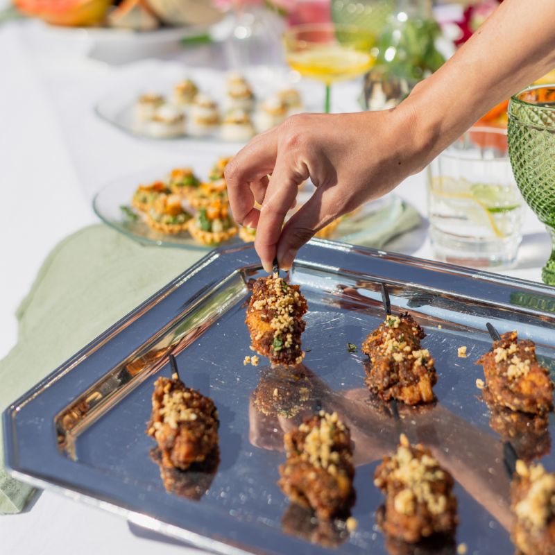 Une cheffe souriante en tenue blanche dresse une table élégante en extérieur avec des amuse-bouches, des plats colorés et des compositions florales, illustrant une expérience traiteur printanière raffinée et conviviale.