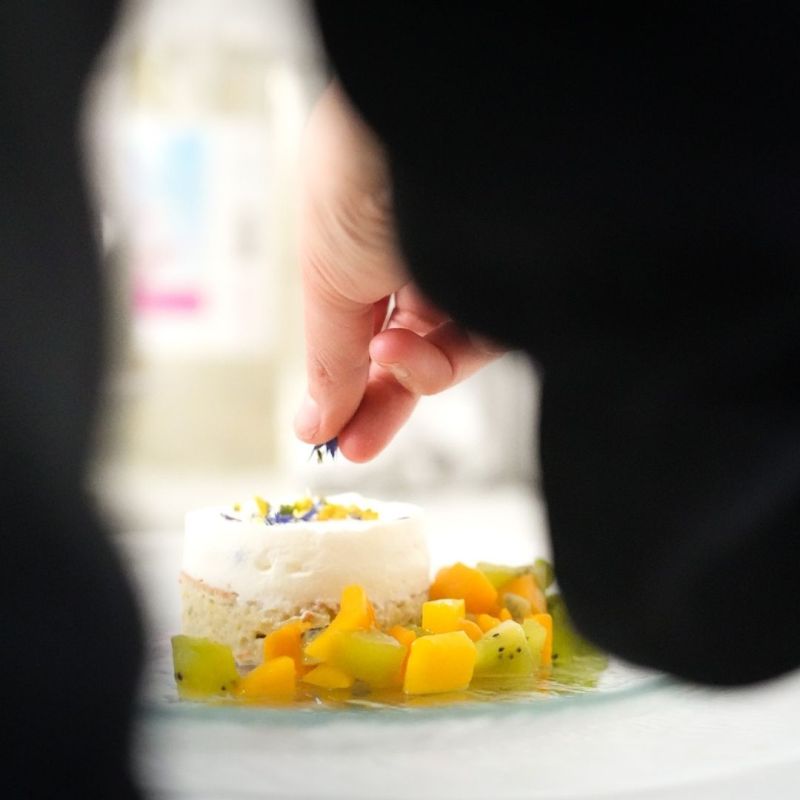Une main verse une boisson rose rafraîchissante dans un verre sur une table élégante en extérieur, entourée de fruits, de fleurs et de plats colorés, illustrant une ambiance conviviale et estivale.