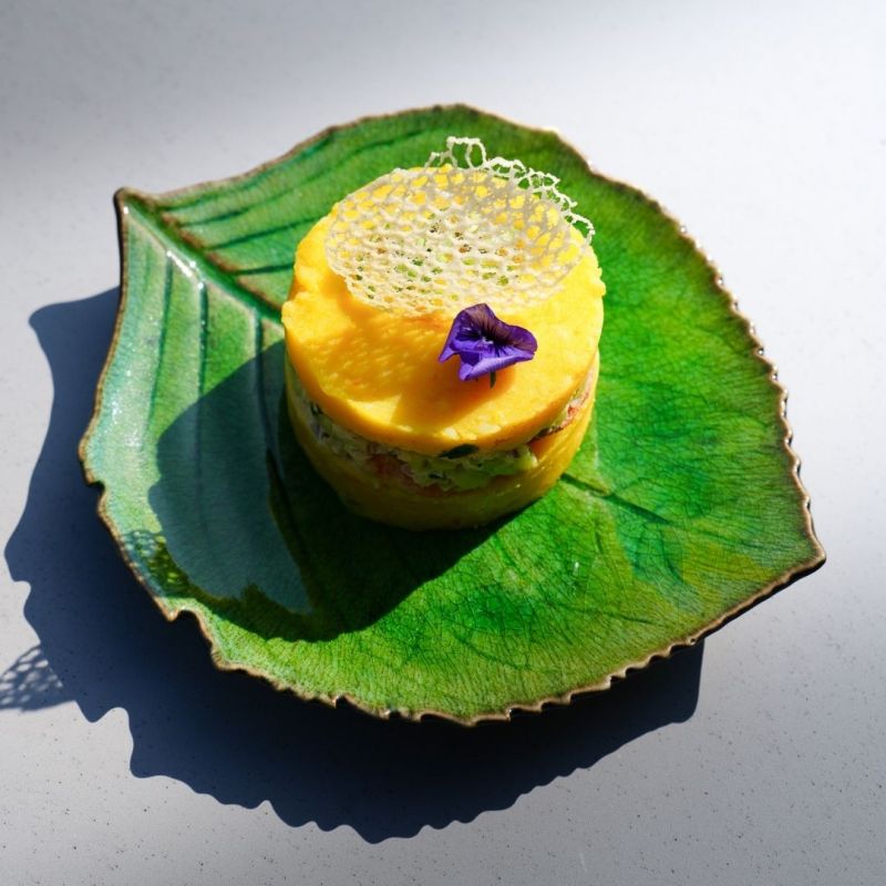 Une cheffe souriante en tenue blanche dresse une table élégante en extérieur avec des amuse-bouches, des plats colorés et des compositions florales, illustrant une expérience traiteur printanière raffinée et conviviale.