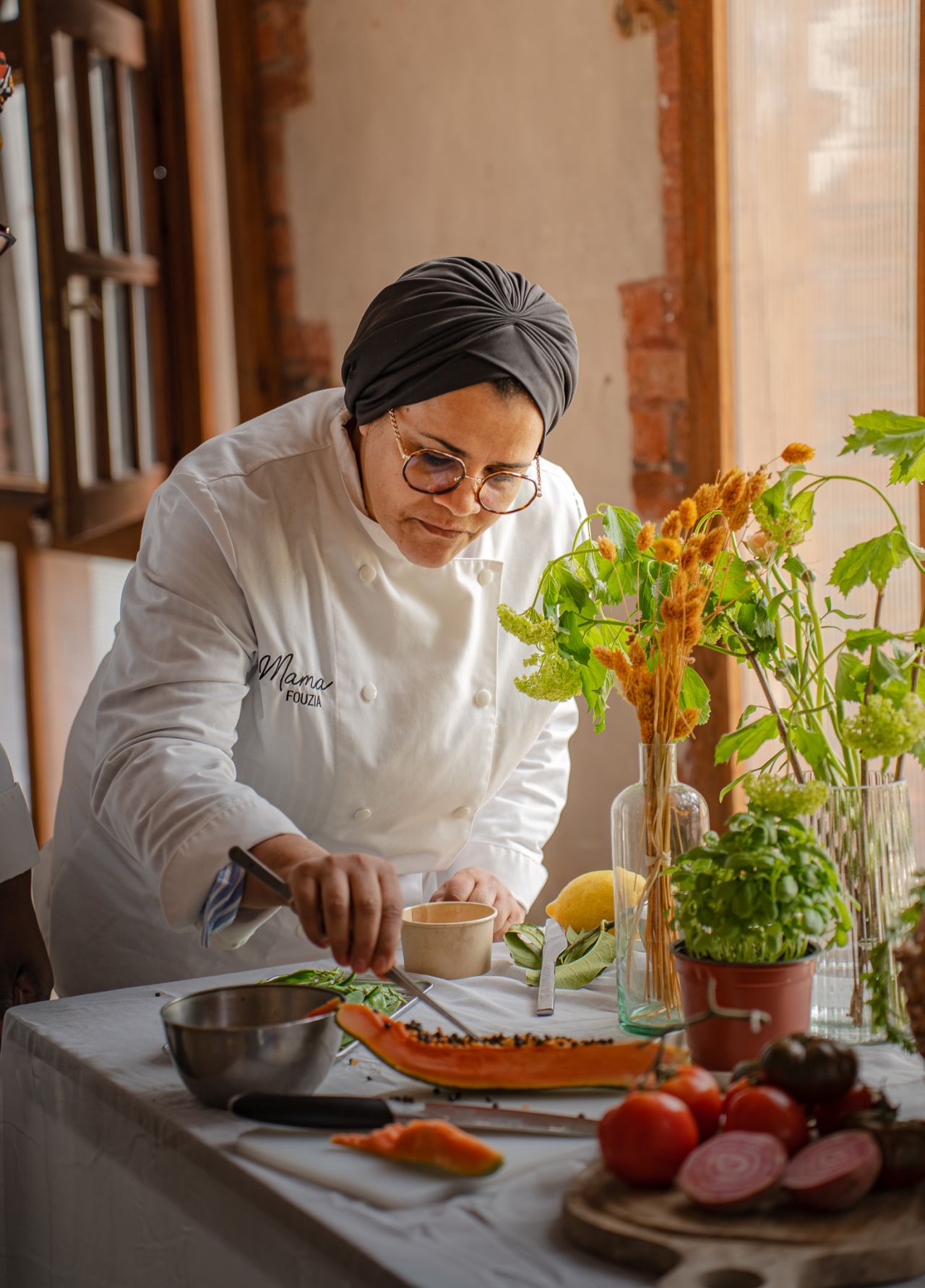 Une main verse une boisson rose rafraîchissante dans un verre sur une table élégante en extérieur, entourée de fruits, de fleurs et de plats colorés, illustrant une ambiance conviviale et estivale.
