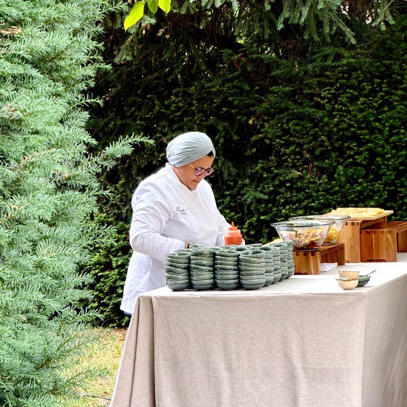 Une cheffe souriante en tenue blanche dresse une table élégante en extérieur avec des amuse-bouches, des plats colorés et des compositions florales, illustrant une expérience traiteur printanière raffinée et conviviale.