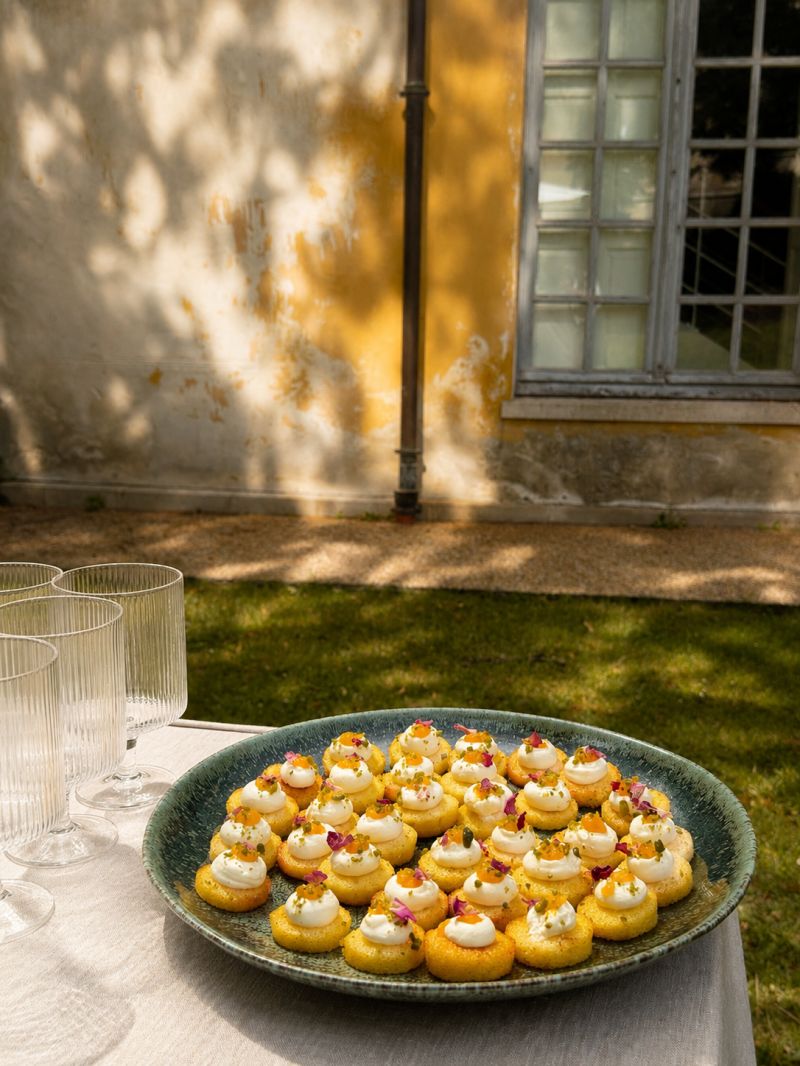 Une main verse une boisson rose rafraîchissante dans un verre sur une table élégante en extérieur, entourée de fruits, de fleurs et de plats colorés, illustrant une ambiance conviviale et estivale.