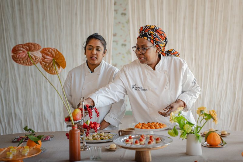 Une table extérieure élégante garnie de fruits frais, légumes de saison, pastèque, agrumes et bouchées apéritives, accompagnée de compositions florales colorées, illustrant une expérience traiteur printanière fraîche et conviviale.