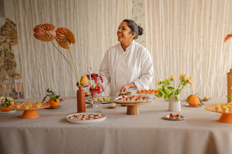 Une cheffe souriante en tenue blanche dresse une table élégante en extérieur avec des amuse-bouches, des plats colorés et des compositions florales, illustrant une expérience traiteur printanière raffinée et conviviale.