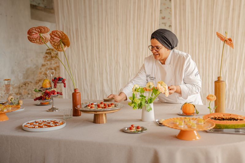 Une main verse une boisson rose rafraîchissante dans un verre sur une table élégante en extérieur, entourée de fruits, de fleurs et de plats colorés, illustrant une ambiance conviviale et estivale.