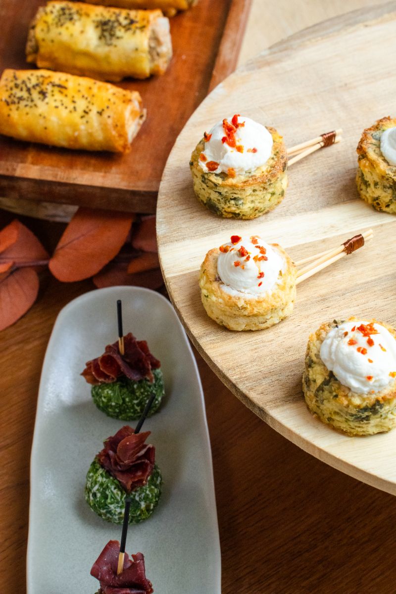 Une cheffe souriante en tenue blanche dresse une table élégante en extérieur avec des amuse-bouches, des plats colorés et des compositions florales, illustrant une expérience traiteur printanière raffinée et conviviale.