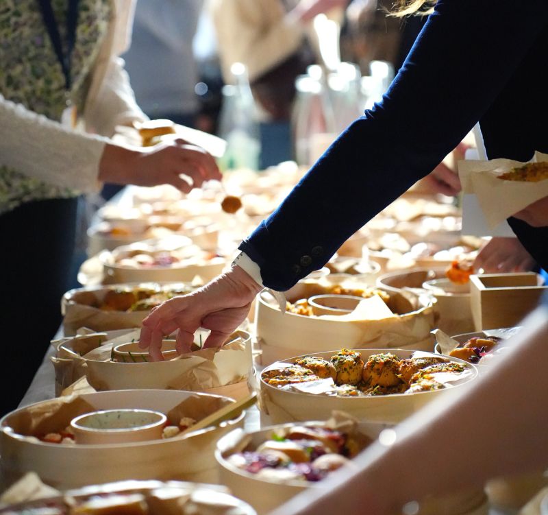 Une table extérieure élégante garnie de fruits frais, légumes de saison, pastèque, agrumes et bouchées apéritives, accompagnée de compositions florales colorées, illustrant une expérience traiteur printanière fraîche et conviviale.