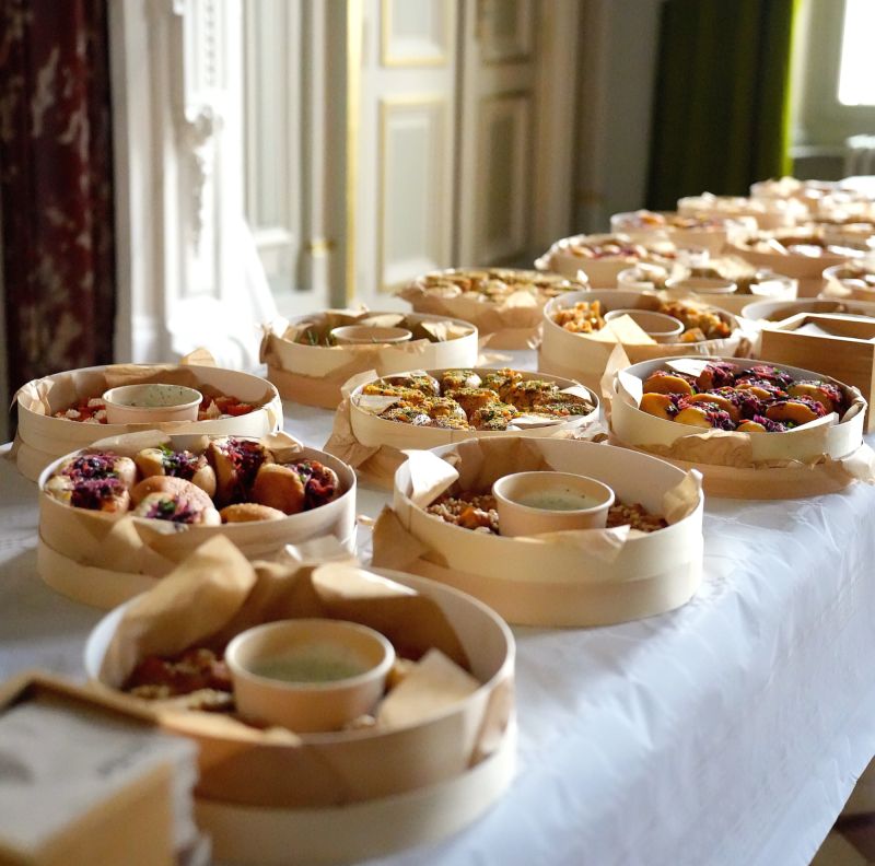 Une cheffe souriante en tenue blanche dresse une table élégante en extérieur avec des amuse-bouches, des plats colorés et des compositions florales, illustrant une expérience traiteur printanière raffinée et conviviale.