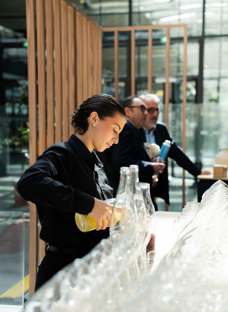 Une cheffe souriante en tenue blanche dresse une table élégante en extérieur avec des amuse-bouches, des plats colorés et des compositions florales, illustrant une expérience traiteur printanière raffinée et conviviale.