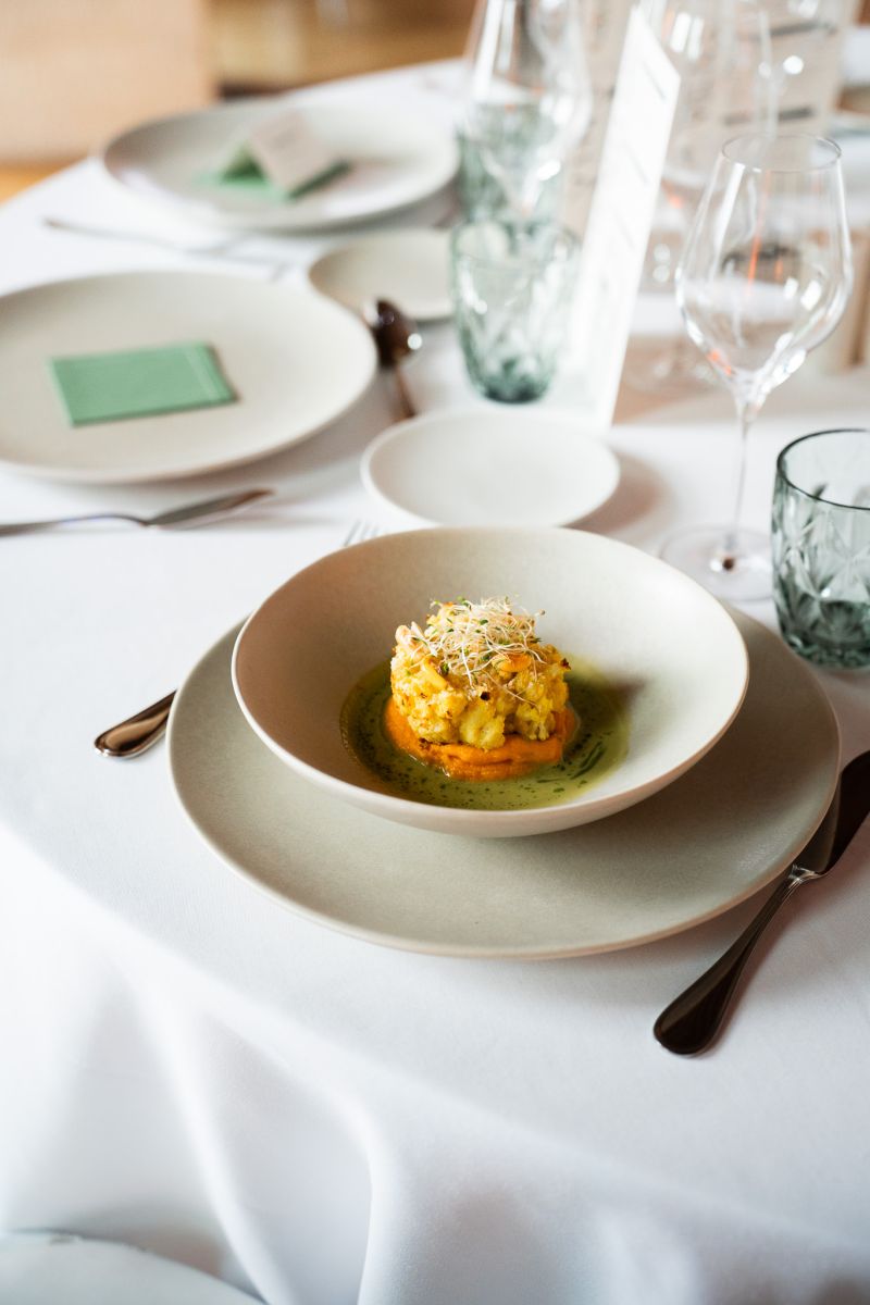 Une cheffe souriante en tenue blanche dresse une table élégante en extérieur avec des amuse-bouches, des plats colorés et des compositions florales, illustrant une expérience traiteur printanière raffinée et conviviale.