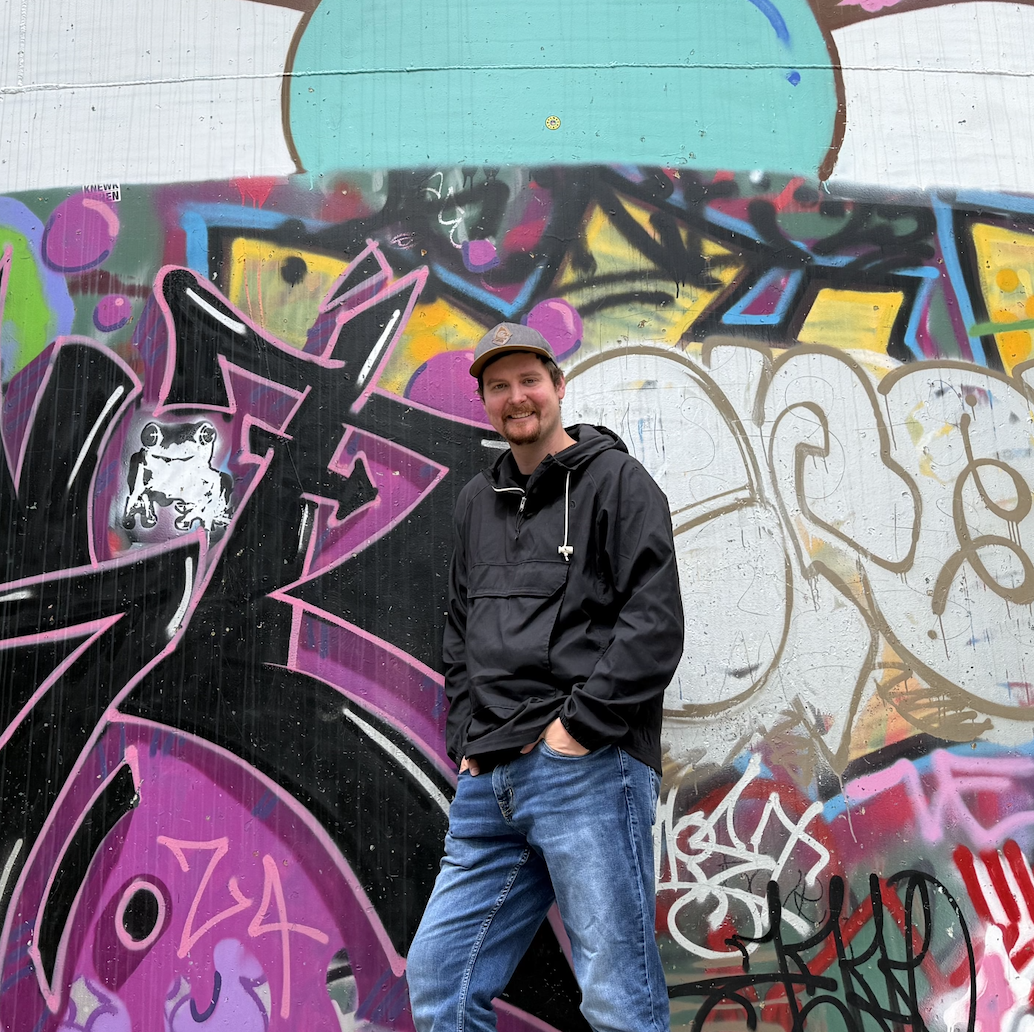 Caleb Pendley smiling in a black shirt and jeans, leaning against a graffiti wall.