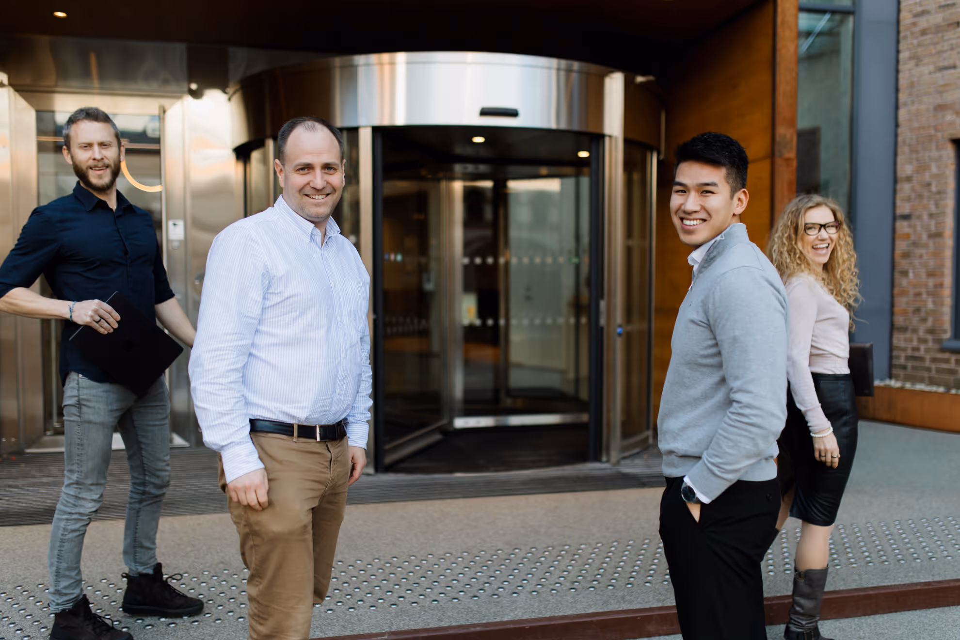 Four diverse business professionals smiling and standing outside an office building with a revolving door.