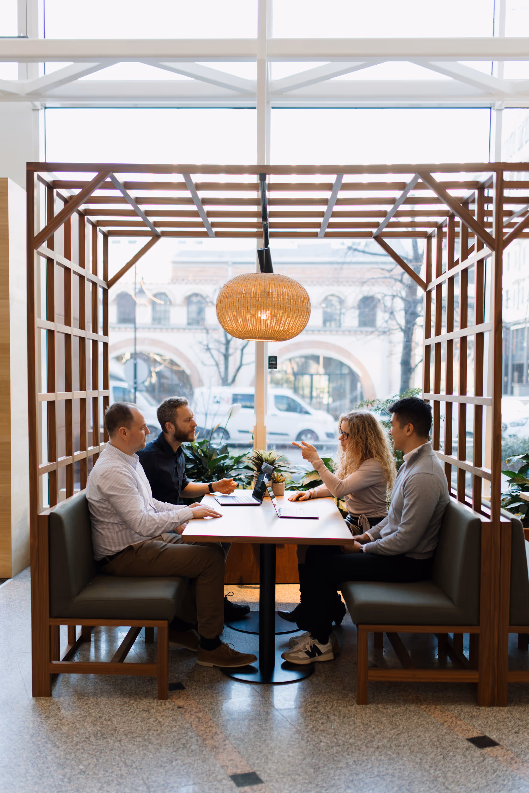 Four people sitting across from each other at a wooden table in a modern indoor booth, engaged in discussion with laptops open.
