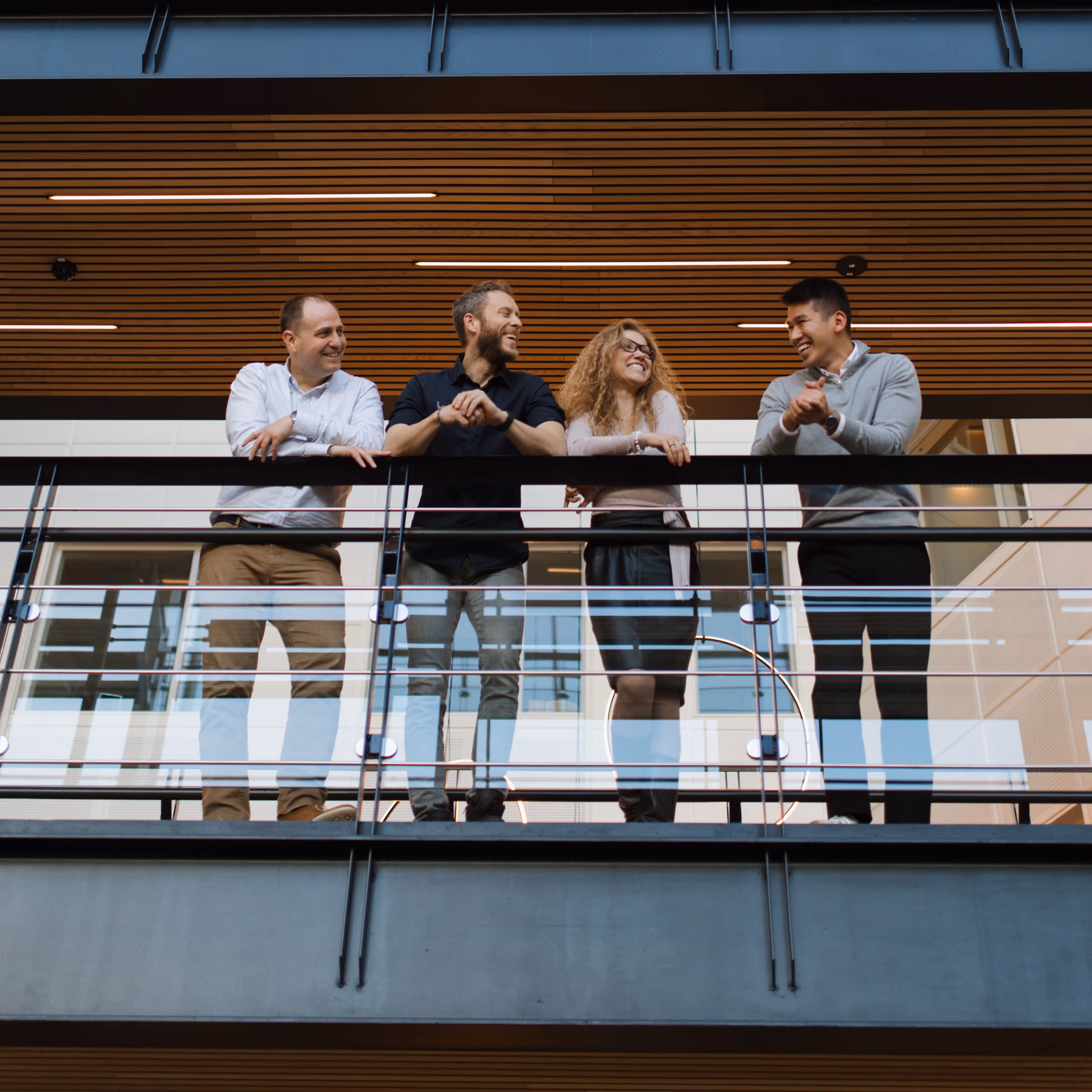 Four diverse colleagues smiling and leaning on a glass railing in a modern office building.