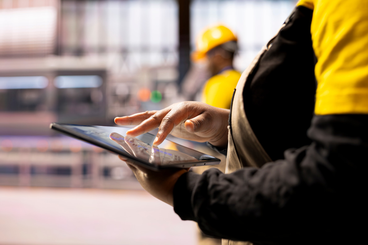 Close-up of a person using a digital tablet in a construction environment with a worker wearing a yellow helmet in the background.