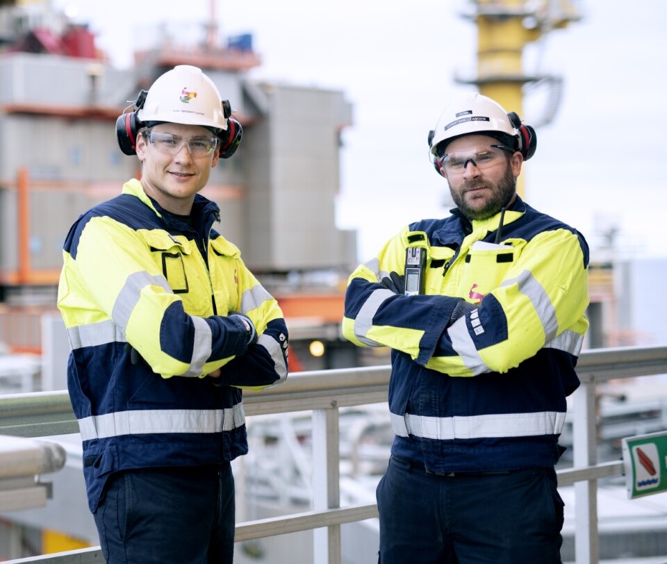 Two industrial workers in yellow and navy safety jackets and white helmets standing with arms crossed at an outdoor worksite.