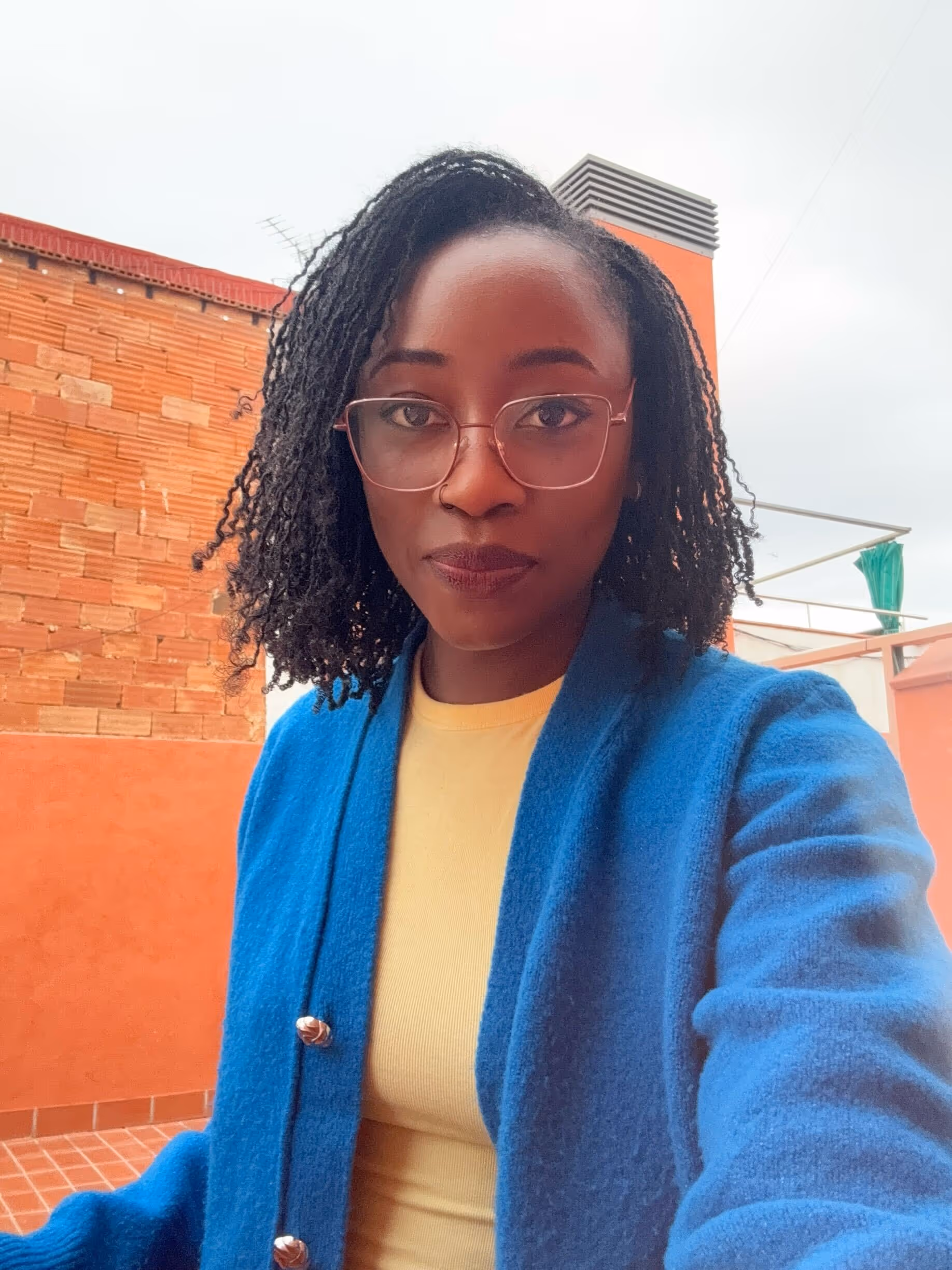 Woman with glasses and shoulder-length curly hair wearing a blue coat and yellow top in an outdoor setting with orange brick walls.