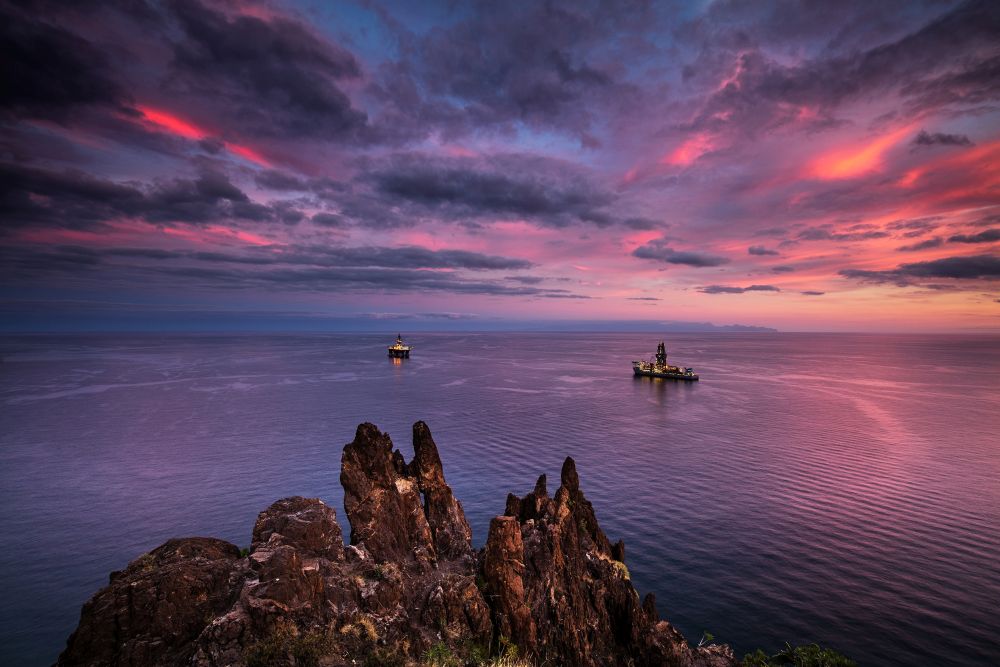 Two illuminated offshore drilling rigs on calm sea waters at sunset with rocky cliffs in the foreground and colorful clouds in the sky.