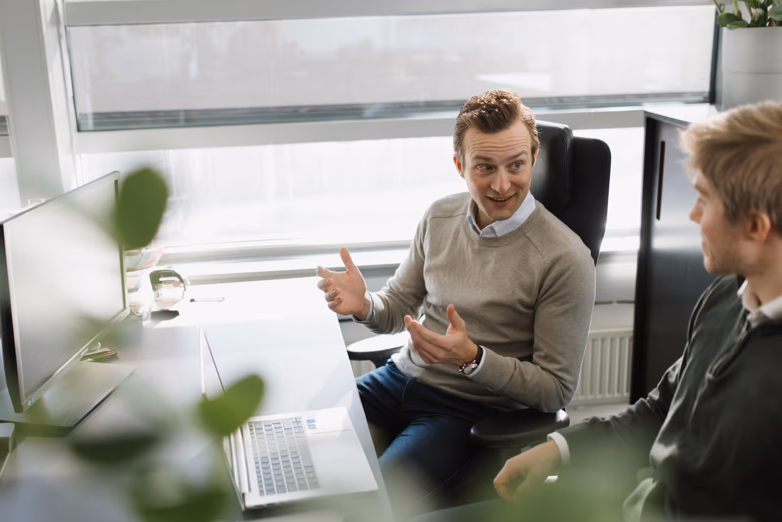 Two men sitting at a desk in a bright office having a conversation with a laptop and monitor in front of them.