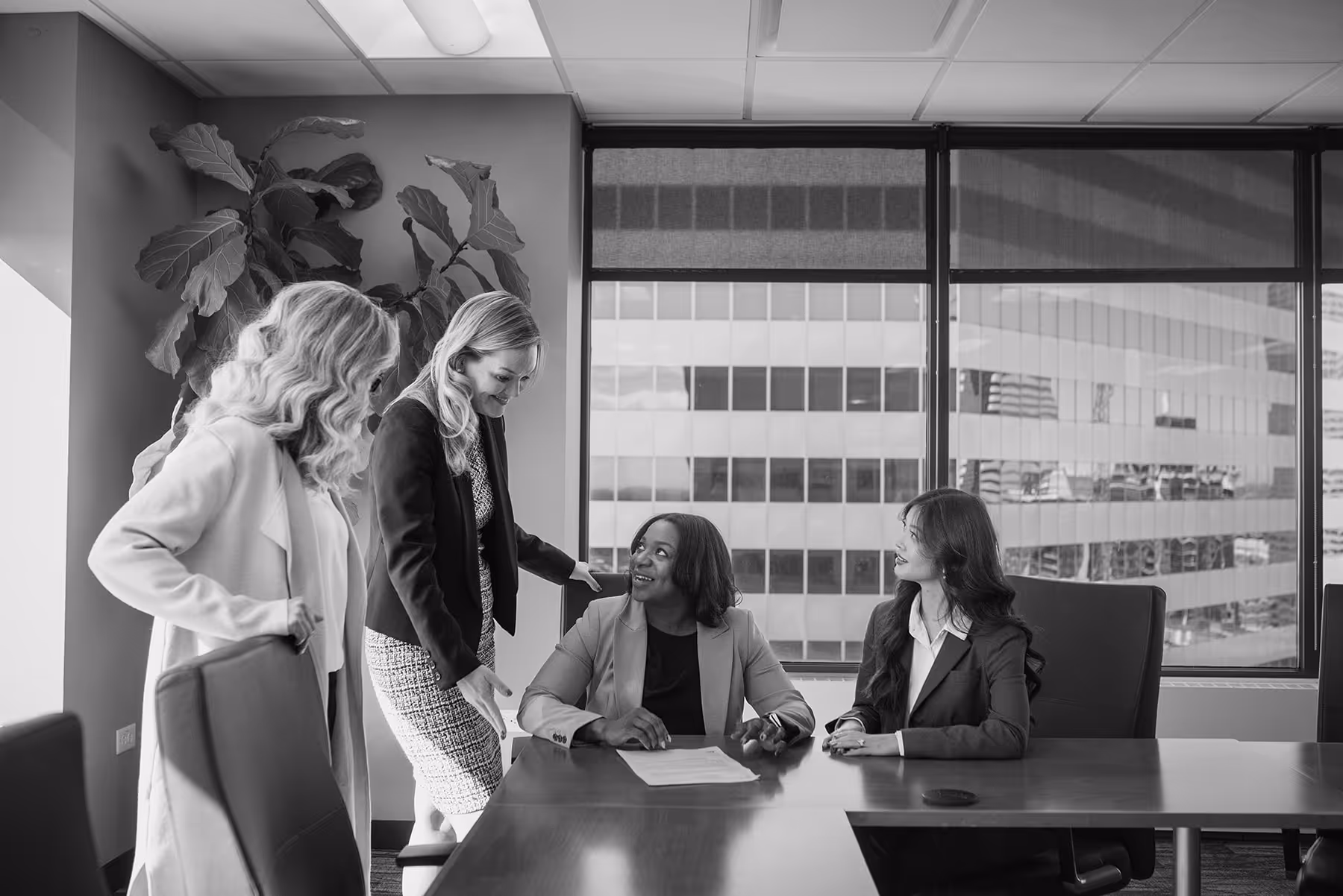 Four businesswomen in a meeting room, two standing and two seated at a conference table with city buildings visible through large windows.