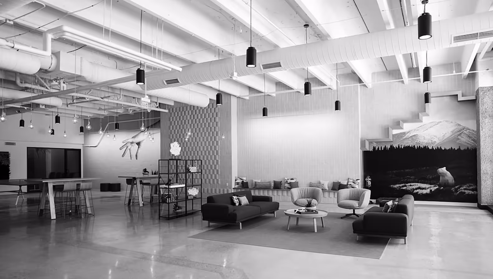 Modern open office lounge with sofas, armchairs, round coffee table, long bench with cushions, and decorative mountain and bear mural.