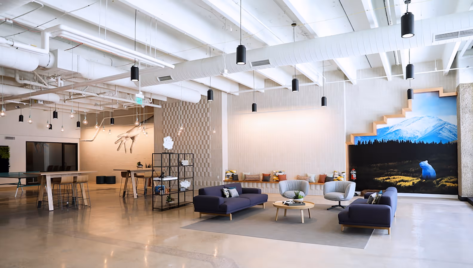 Modern open lounge area with blue sofas, gray armchairs, a round wooden coffee table, pendant lights, and a mural of a bear with mountains in the background.
