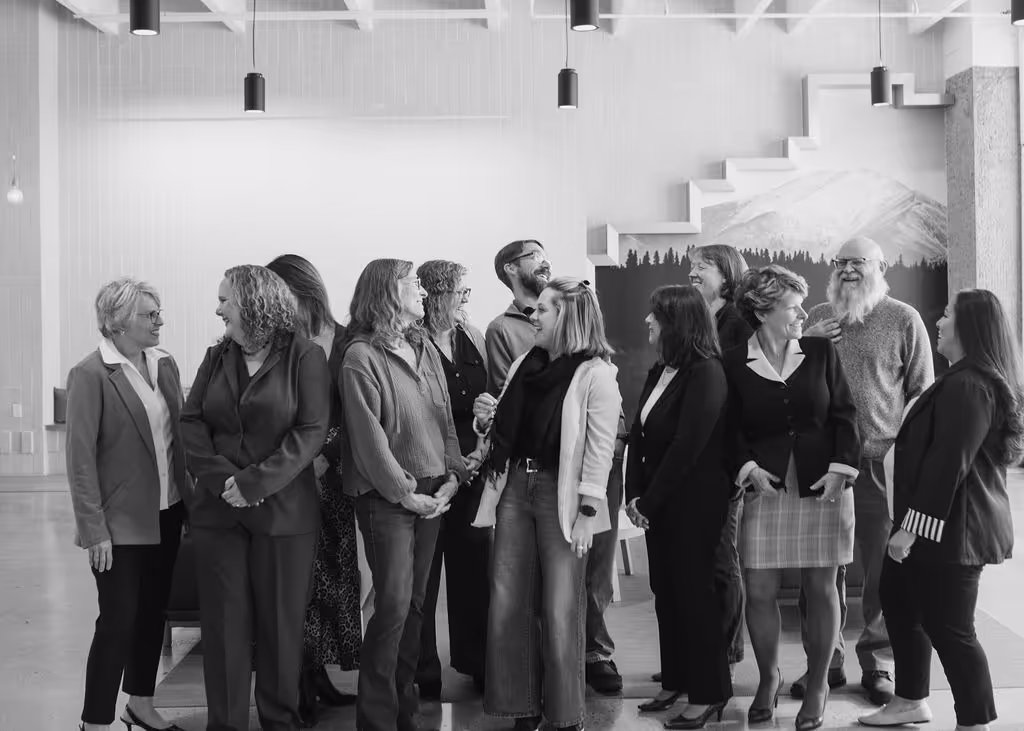 Group of twelve diverse adults standing indoors in a conversation, smiling and interacting in a modern room with a large wall mural of a mountain and forest.