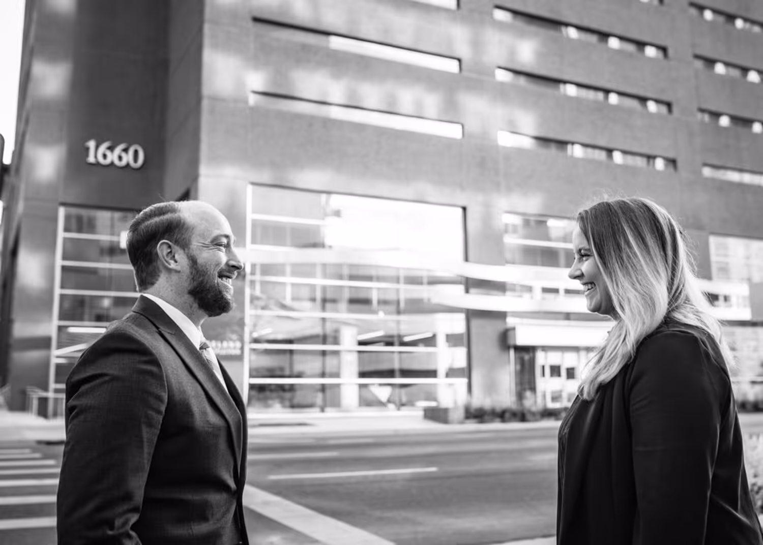 Man and woman in business attire conversing on a city sidewalk in front of a modern building.