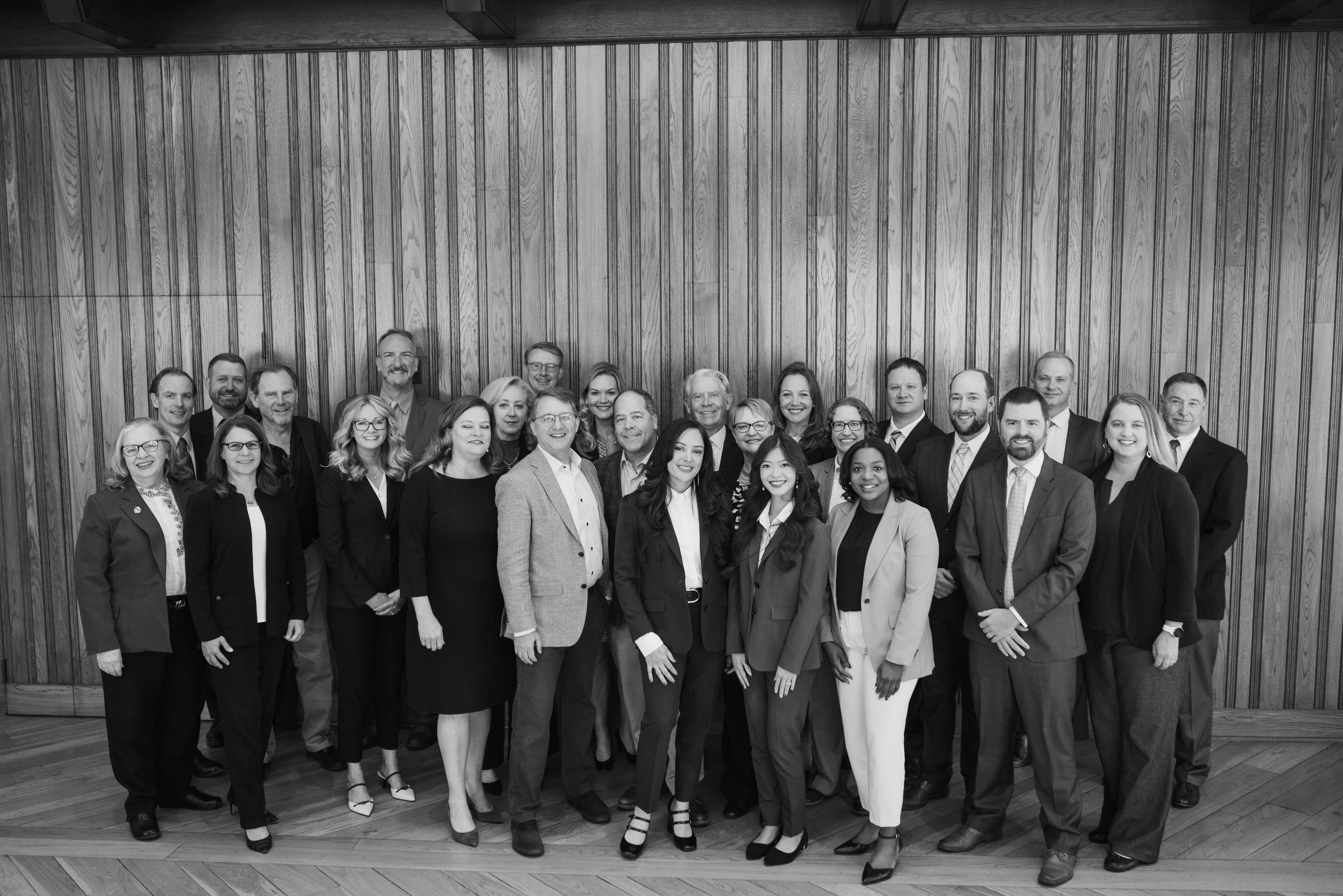 Group portrait of 25 professionally dressed adults standing in front of a vertical wooden panel wall.
