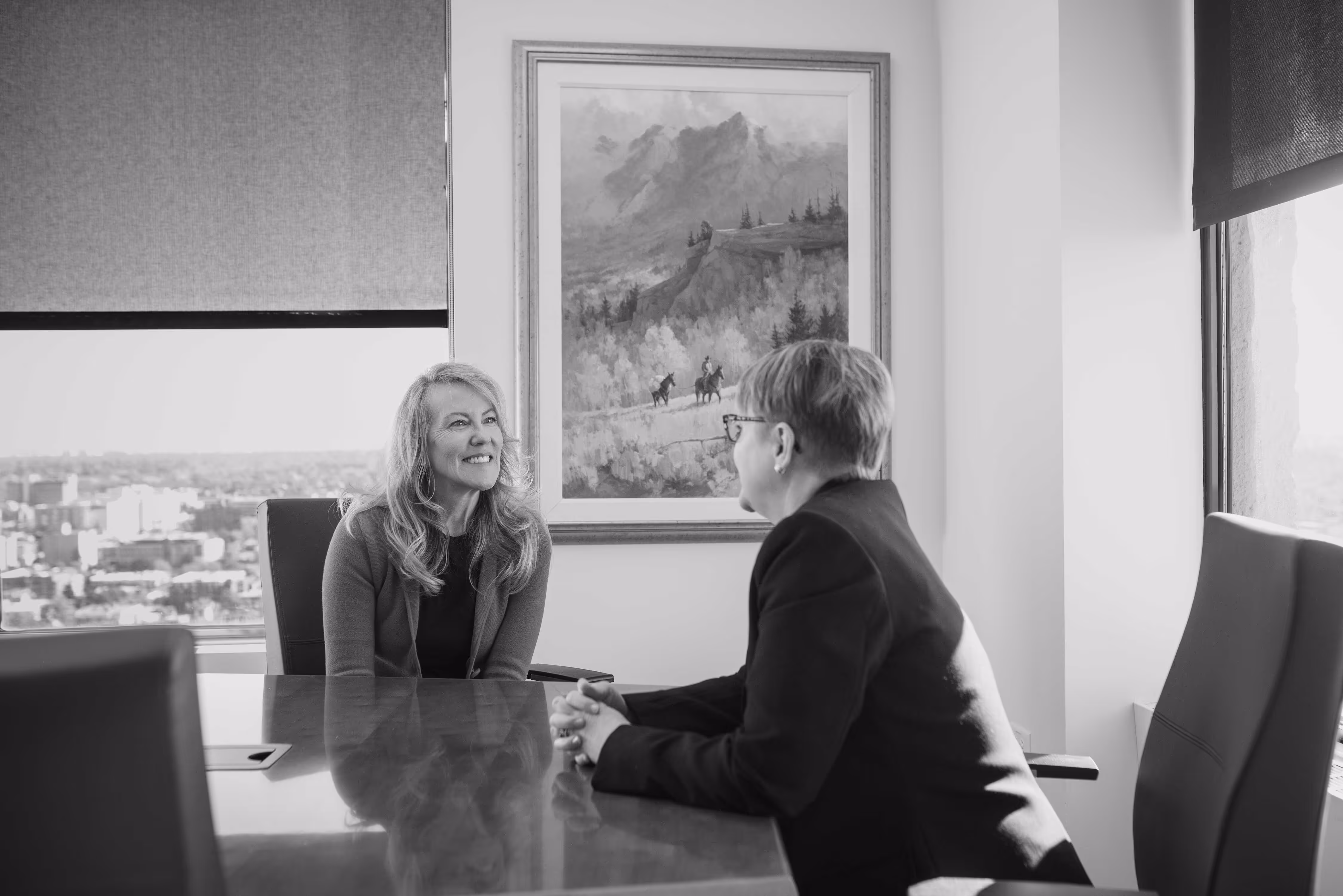 Two women having a conversation across a conference table in an office with city view and a framed mountain painting on the wall.