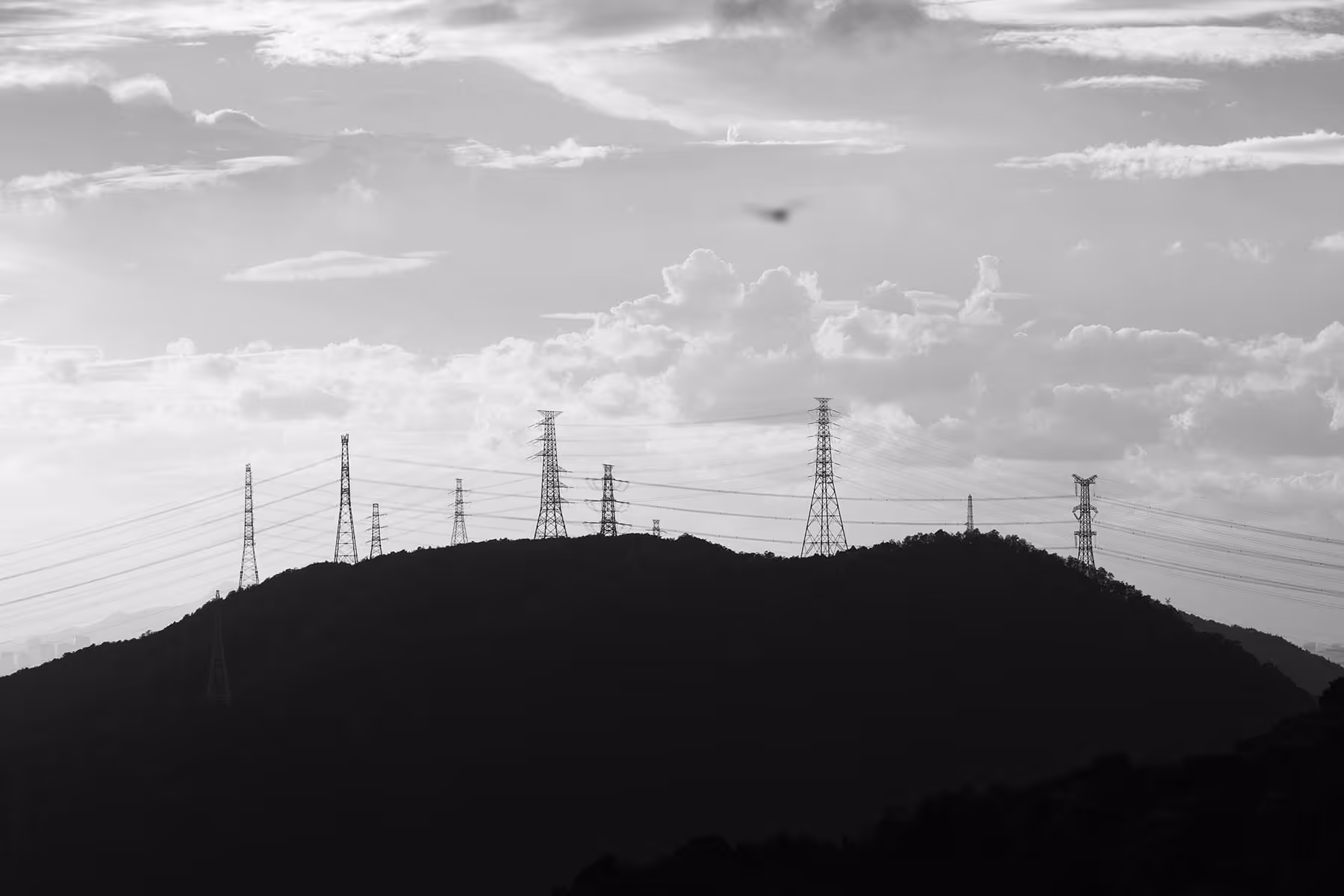 Silhouetted mountain ridge with multiple electricity transmission towers and power lines against a cloudy sky.