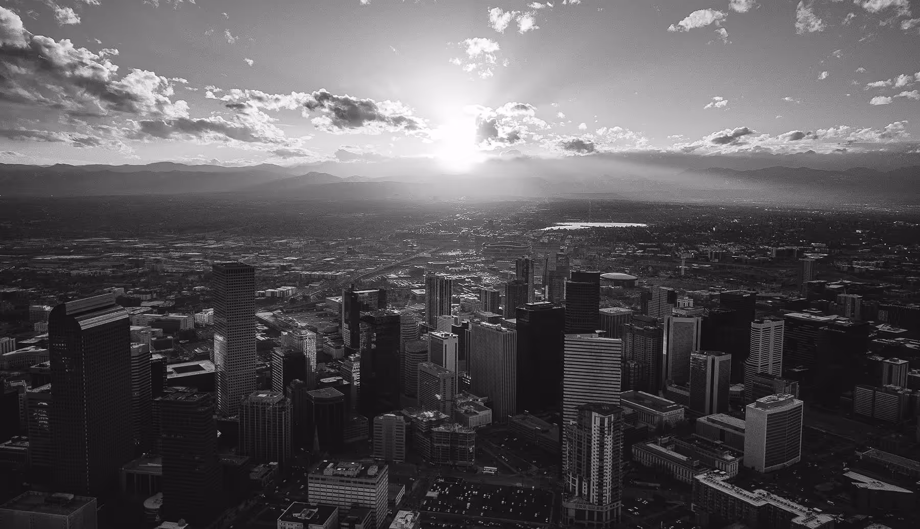 Black and white aerial view of a city skyline with the sun setting behind distant mountains and scattered clouds in the sky.