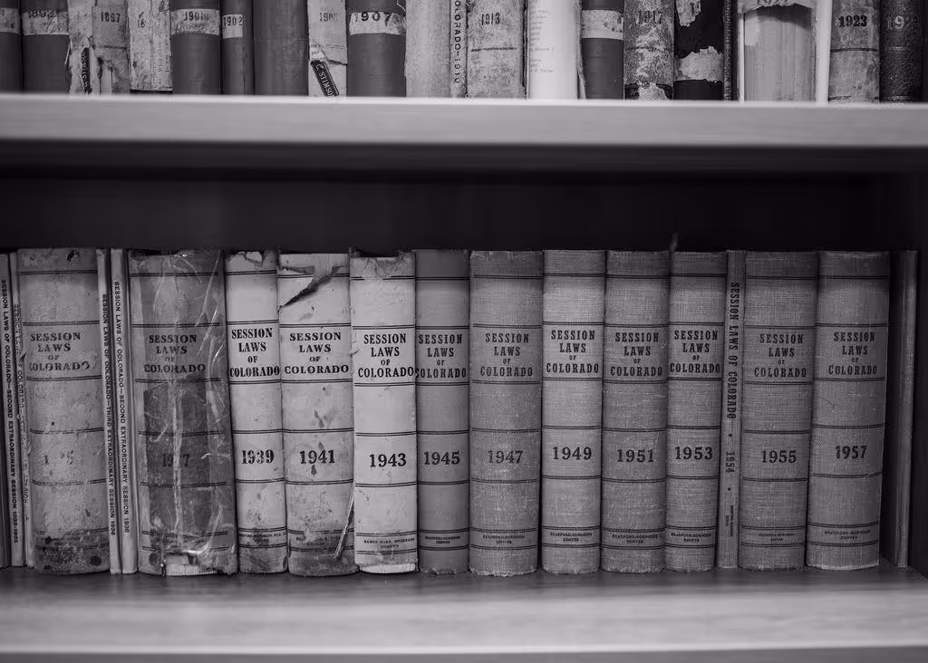Black and white image of a row of old legal books titled 'Session Laws of Colorado' from various years arranged on a wooden shelf.