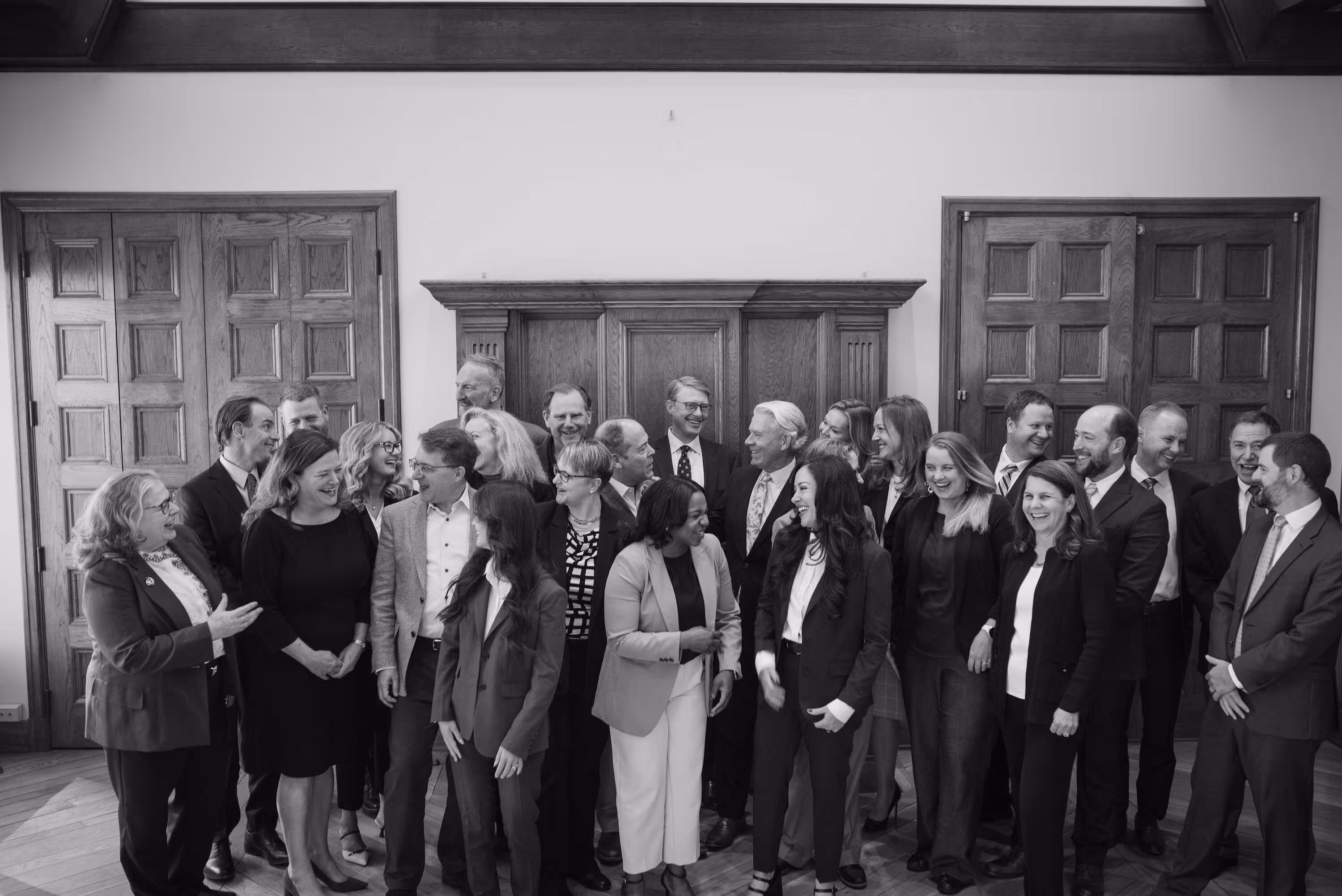 A diverse group of professionally dressed men and women standing indoors, smiling and interacting with each other in front of wooden doors and paneling.