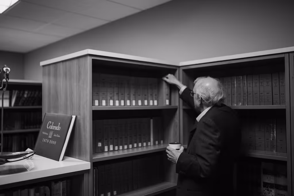 Man in a suit with a cup in hand reaching for a book on a shelf filled with organized books in an office or library.