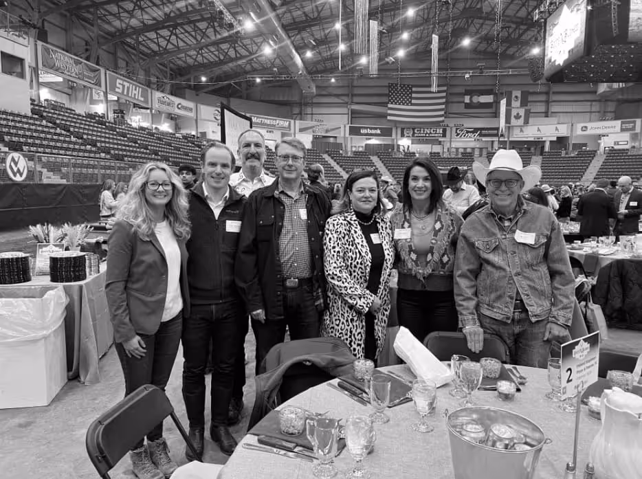 Seven people standing together smiling in an event arena with tables set for dining.