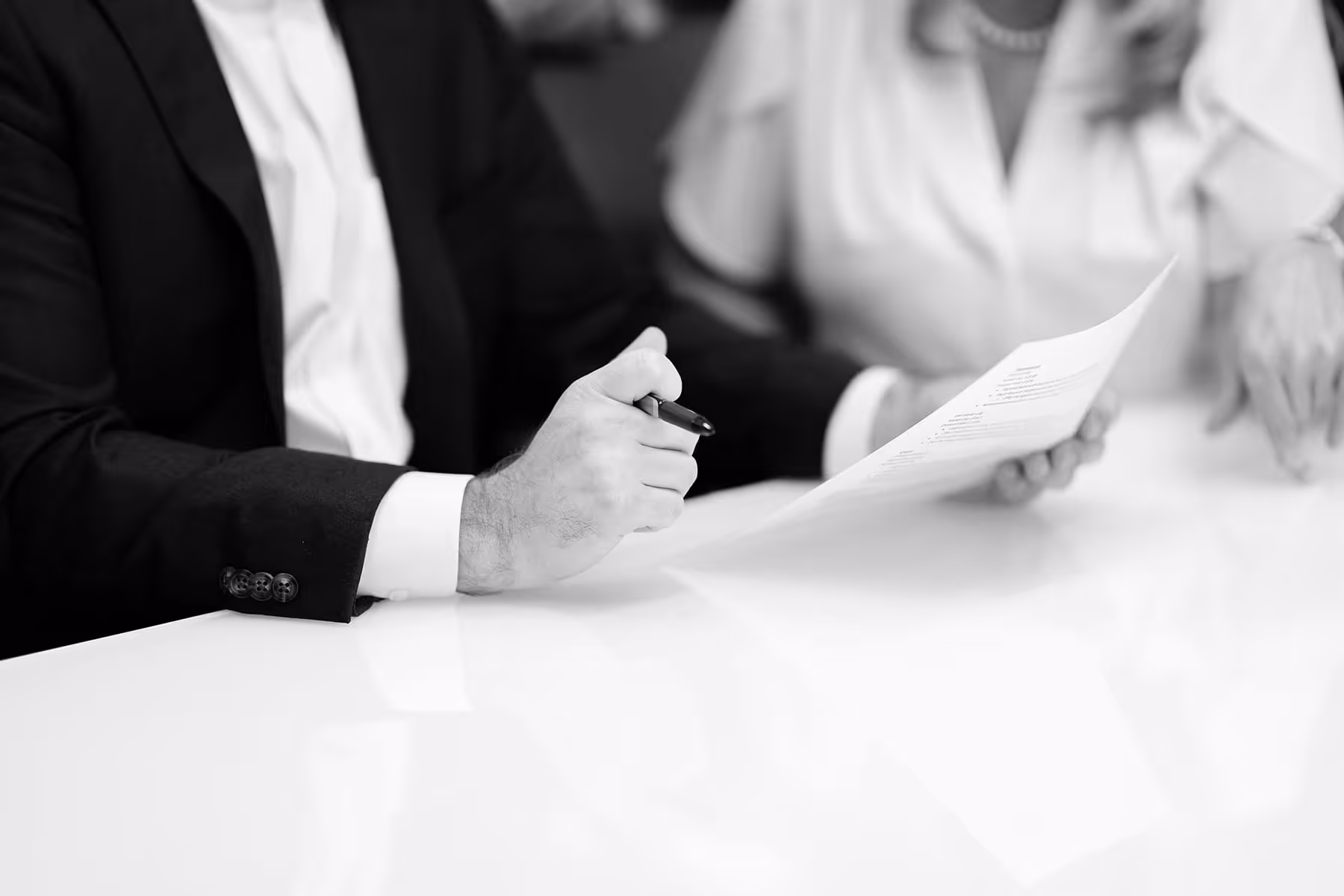 Two people, one in a suit holding a pen and document, sitting at a table in a business setting.