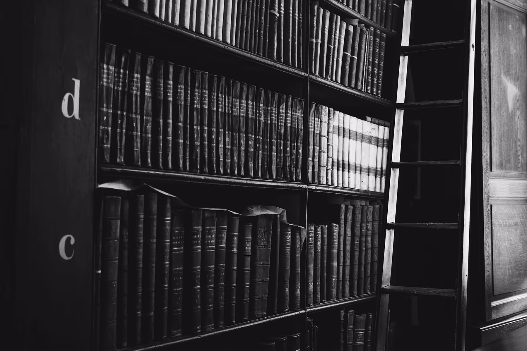 Black and white photo of old leather-bound books on library shelves with a wooden ladder leaning against the shelves.