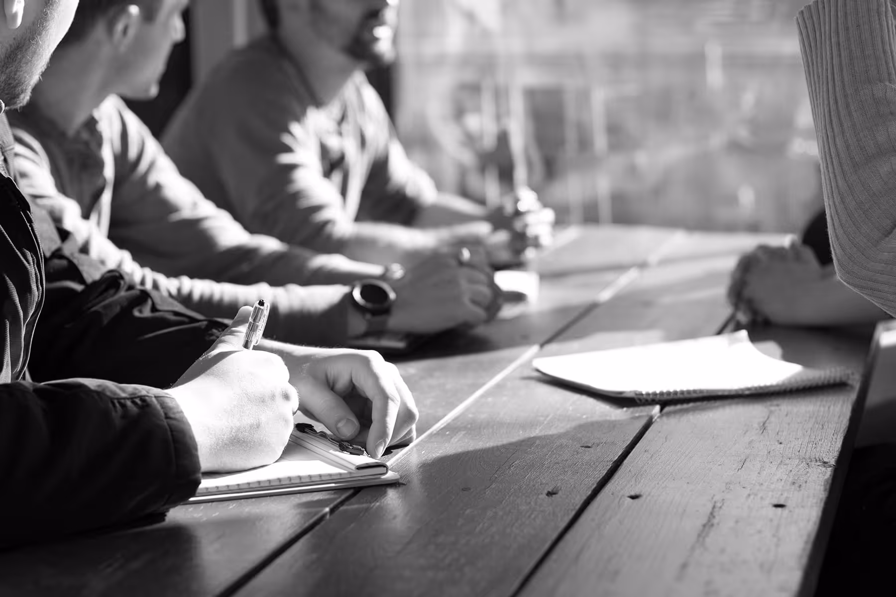 People sitting at a wooden table, writing in notebooks during a meeting.