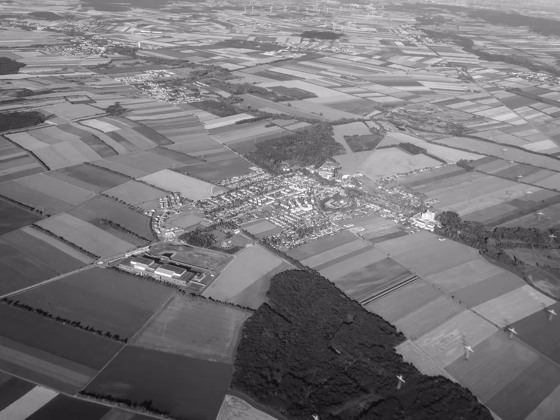Aerial view of a rural landscape with patchwork farmland, small town clusters, wooded areas, and wind turbines in the distance.